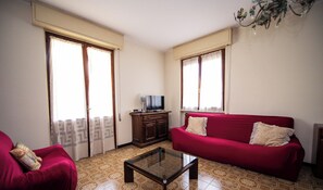 Un salón con un sofá rojo, una mesa de centro de vidrio, un televisor sobre un mueble de madera y baldosas de suelo estampadas.