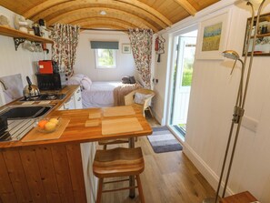A compact kitchen with a sink, kettle, and a fruit bowl, a wooden dining table with a bench, and a cozy bed with a window. - Higher Merther Farm Shepherds Hut (Helston)