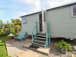 A small, single-story house with a sliding door, a small porch with a bench, and a potted plant. - Higher Merther Farm Shepherds Hut (Helston)