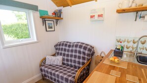 A cozy room with a wooden floor, a checkered sofa, a dining table with a cutting board, a window with a view of greenery, and a shelf with books and a picture. - Higher Merther Farm Shepherds Hut (Helston)