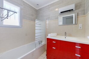 Een badkamer met een bad, een douche, een rode wastafel met spiegel, en een raam.