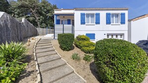 Une maison à deux étages avec un escalier en pierre menant à une terrasse en bois, entourée de haies bien entretenues et sous un ciel dégagé.