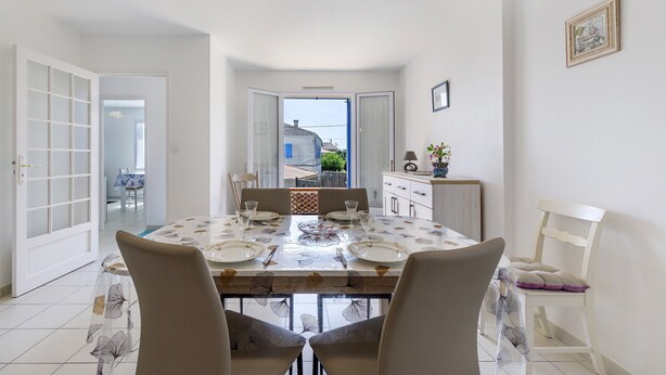Un coin repas avec une table dressée pour quatre personnes, des chaises, une vue sur l’extérieur par une porte ouverte et un petit meuble sur lequel est posée une plante.