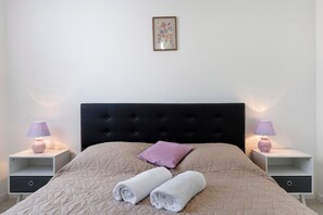 Een net opgemaakt bed met een gewatteerde spreidek, twee nachtkastjes met lampen, een hoofdbord, en een ingelijste foto aan de muur.