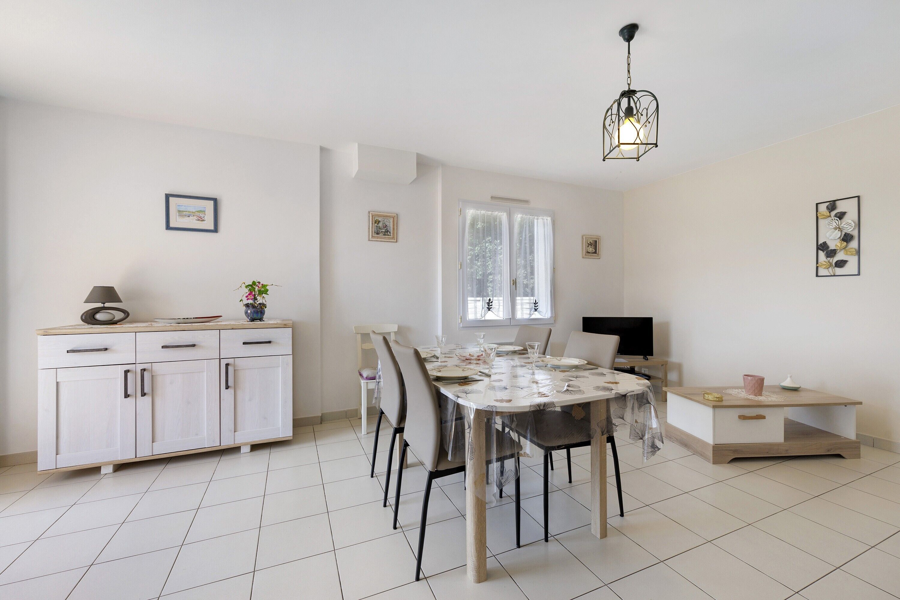 Un comedor con una mesa lista para cuatro, un armario con cajones, un televisor y una ventana con cortinas.