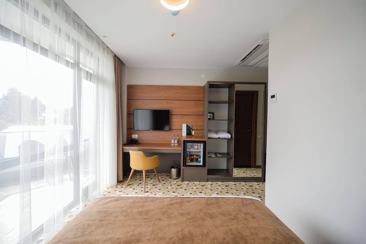 Ett hotellrum med en säng, ett skrivbord, en stol, en TV, en garderob och ett fönster med gardiner.