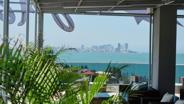 A balcony with wicker furniture, a view of the city skyline, and a clear blue sea. - Calligraphy Hotel (Batumi)