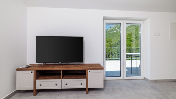 Un salon moderne avec un téléviseur à écran plat posé sur un meuble télé en bois, une porte-fenêtre coulissante donnant sur un balcon et un sol carrelé.