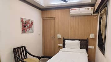 A hotel room with a bed, a chair, a ceiling fan, an air conditioner, and a painting on the wall.