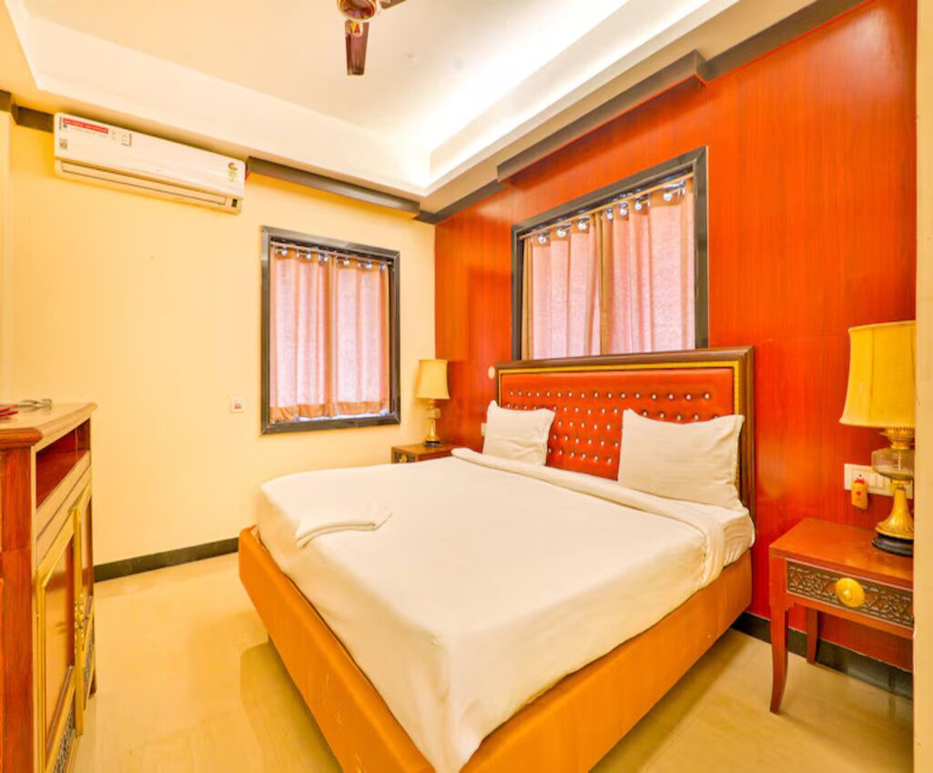 A hotel room with a large bed, two bedside tables with lamps, a window with curtains, and an air conditioning unit.