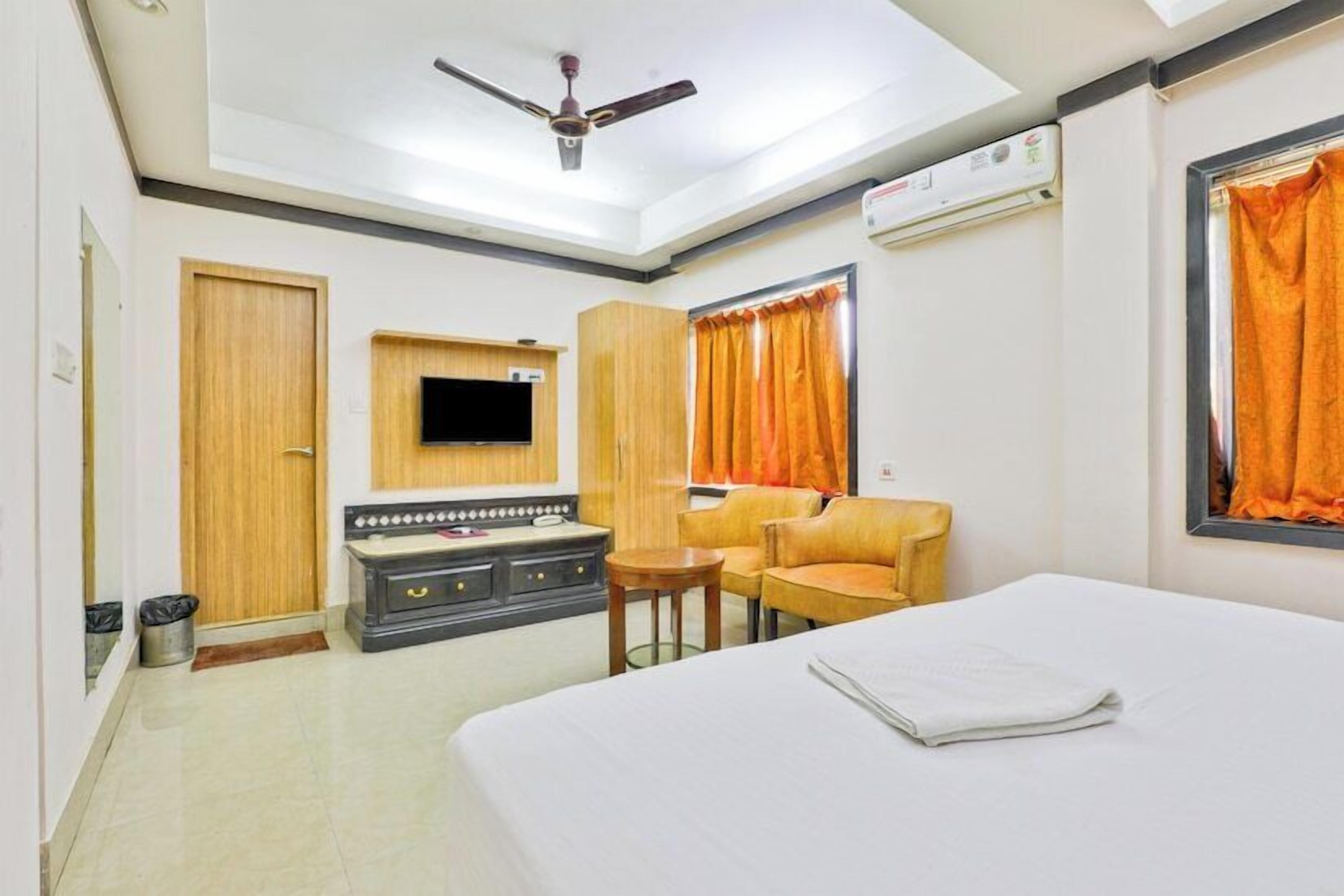 A hotel room with a bed, a TV mounted on the wall, a ceiling fan, an air conditioning unit, and a window with orange curtains.