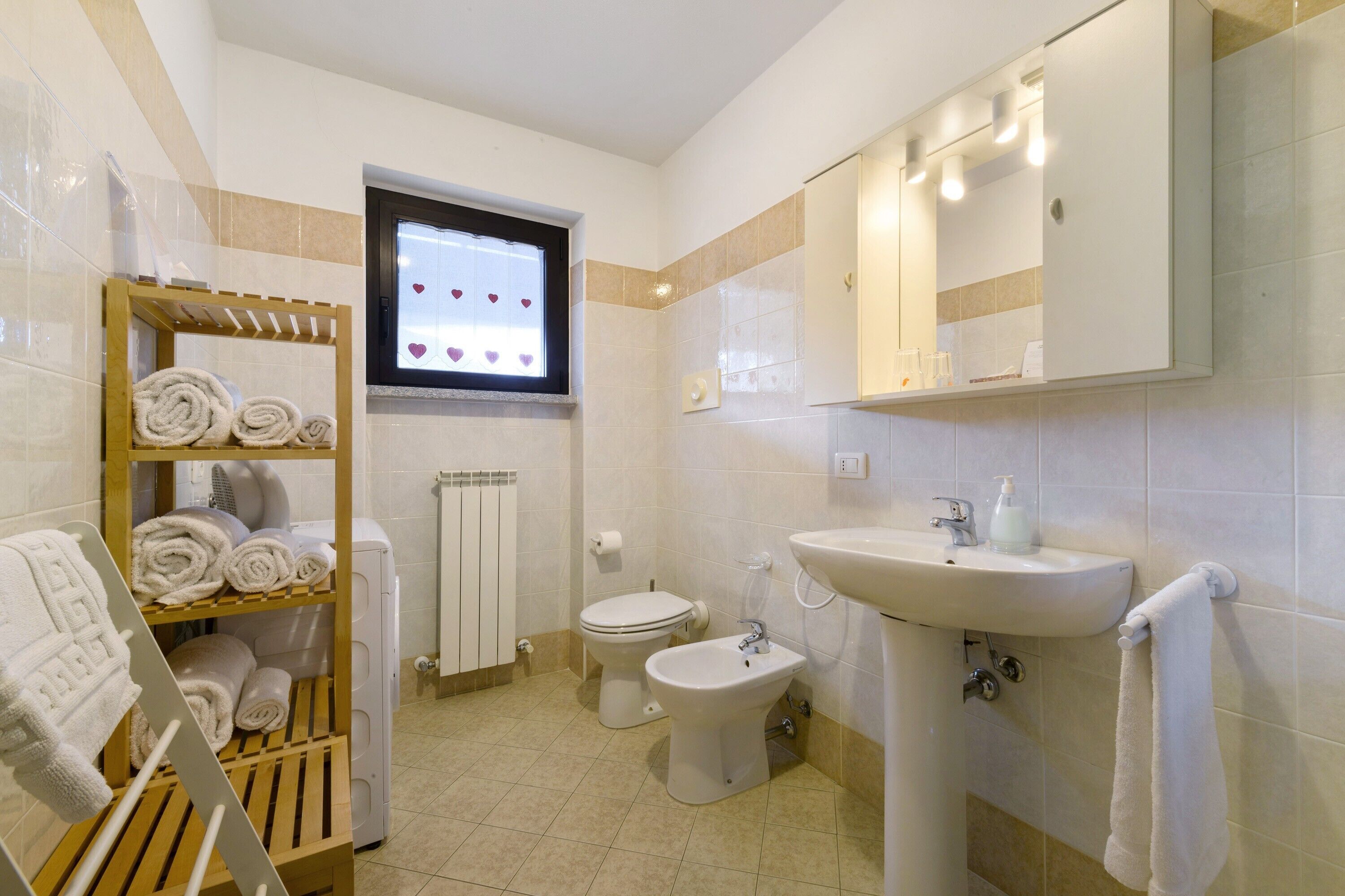 A bathroom with a toilet, bidet, sink, towel rack, and a window with a view of hearts.