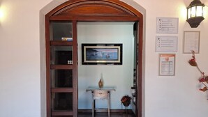 A wooden door with a glass panel, a framed picture on the wall, a small table with a vase, and emergency contact information on the wall. - San Roque village (Posadillo)
