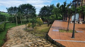 A stone pathway leading to a building with a tiled patio area, surrounded by greenery and trees. - San Roque village (Posadillo)