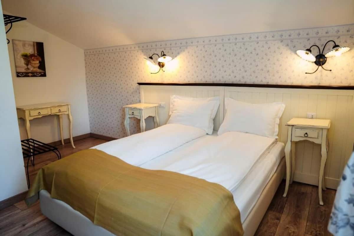 A bedroom with a bed, two bedside tables, a nightstand, a wall-mounted light fixture, and a small table with a chair.