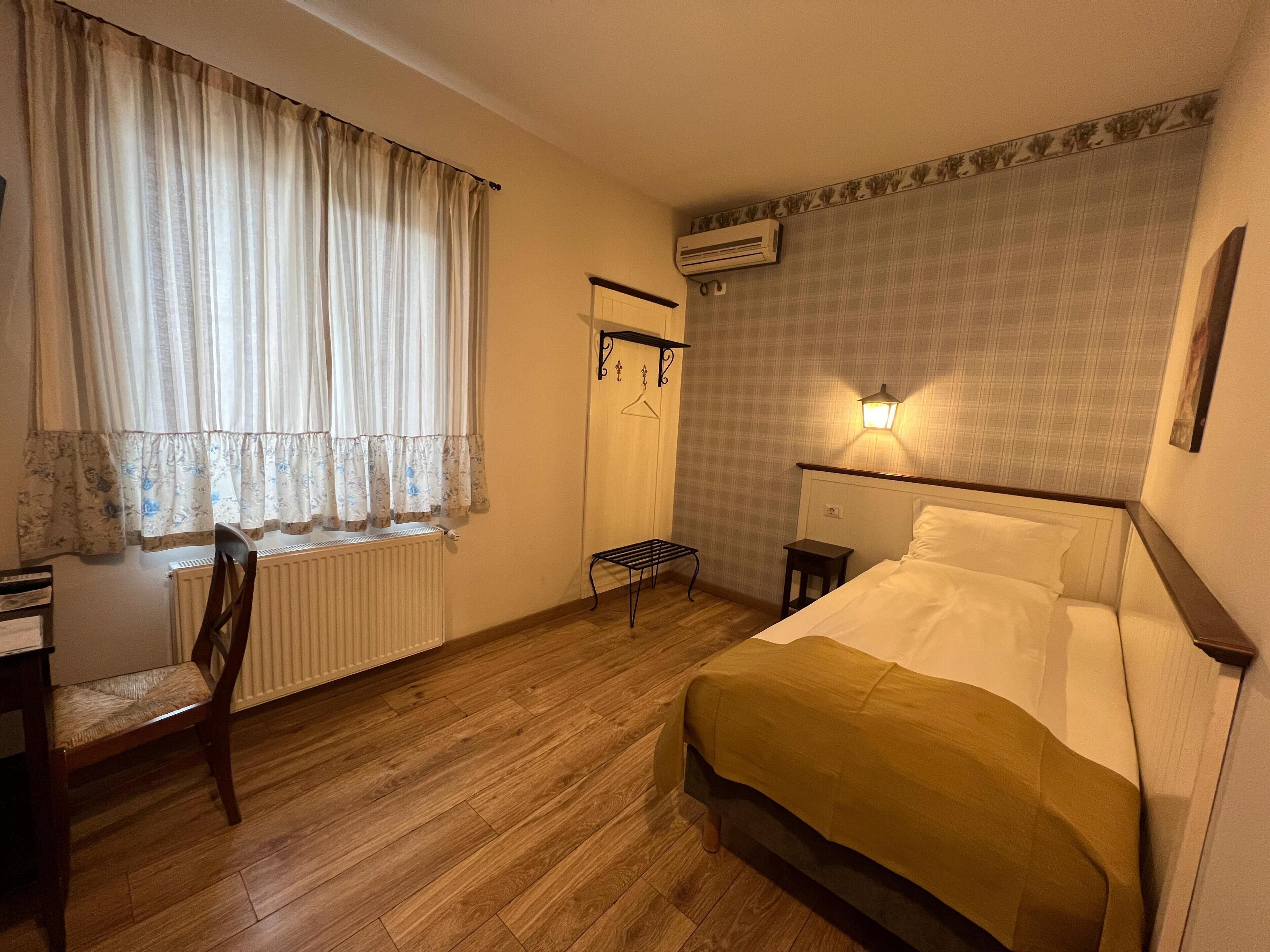 Habitación de hotel con cama, radiador, silla, mesita, perchero y aire acondicionado de pared.