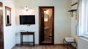 Una stanza con televisione a muro, un tavolino con lampada, una porta che dà su un bagno e una finestra con tende.