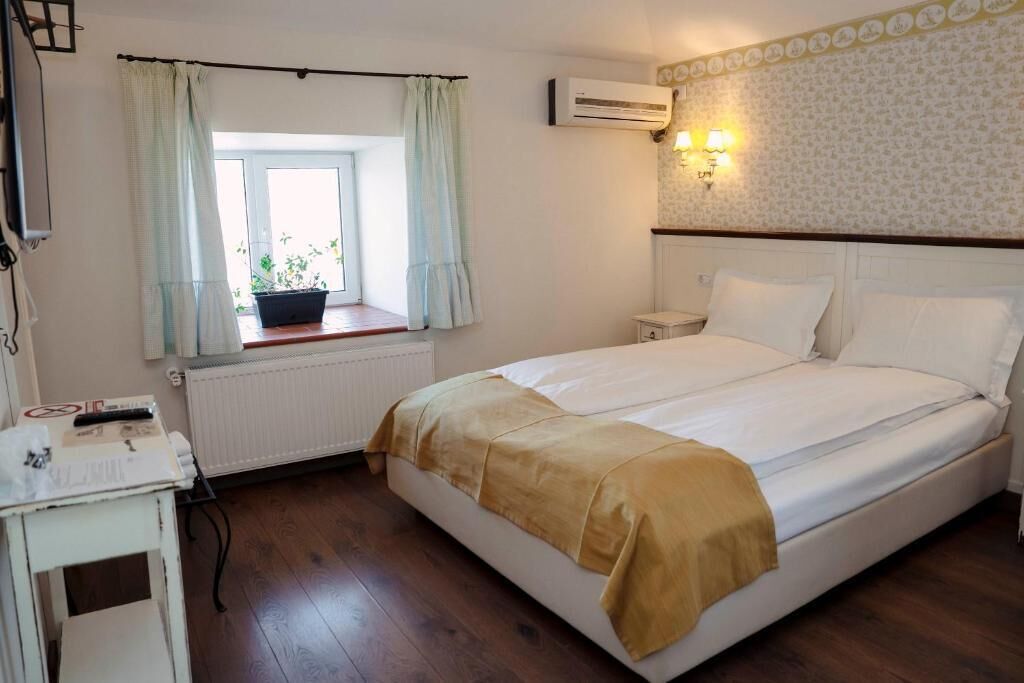 Un dormitorio con cama, ventana con cortinas, mesita de noche, unidad de aire acondicionado y lámpara fijada a la pared.
