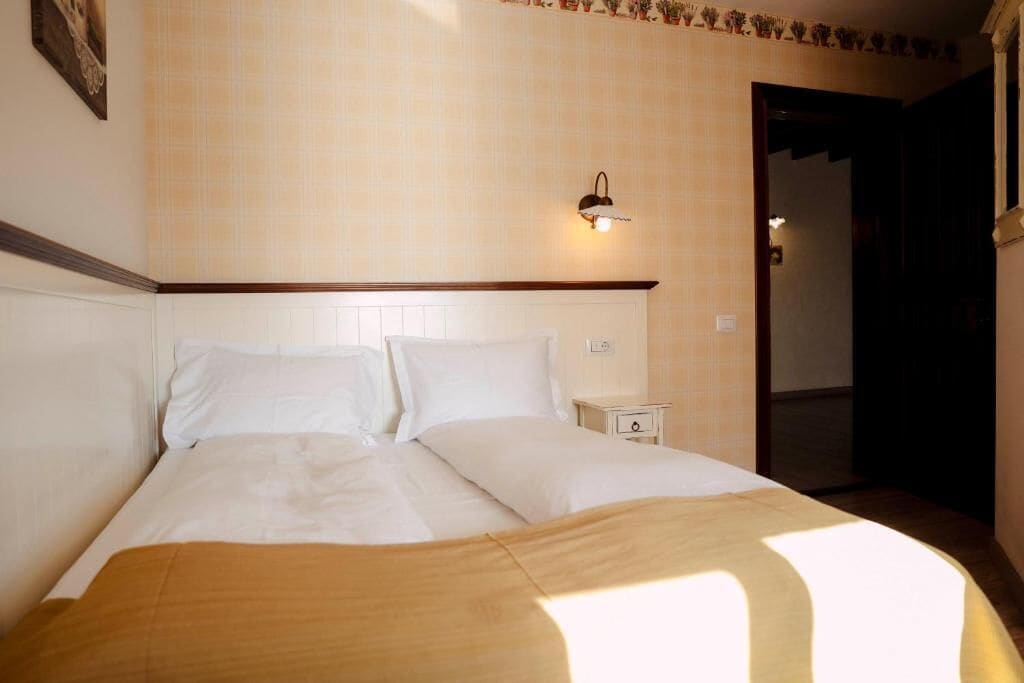 A neatly made bed with white linens, a bedside table with a lamp, and a wall-mounted light fixture.