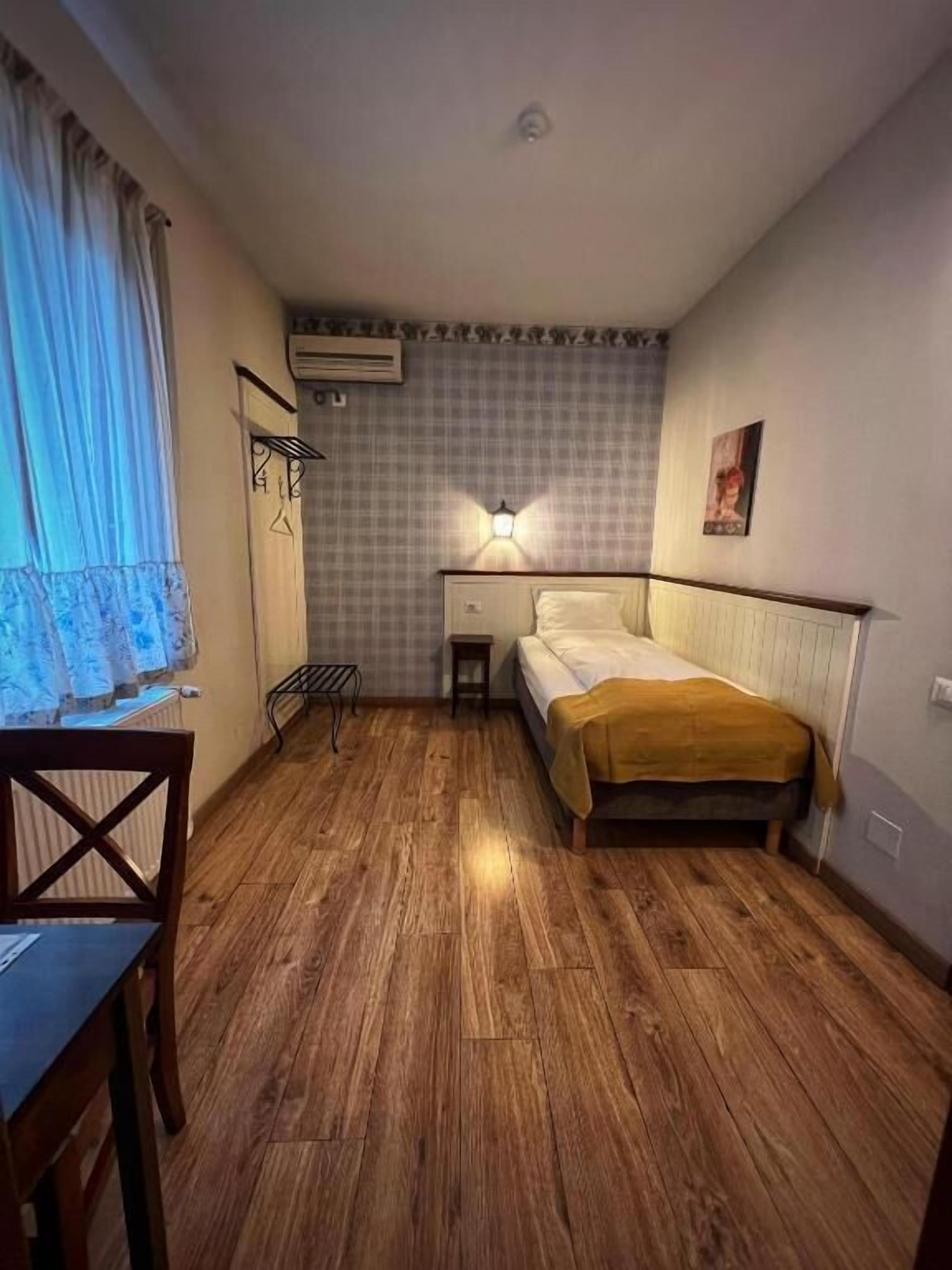 A small, cozy room with a wooden floor, a bed with a yellow blanket, a desk with a chair, and a radiator.