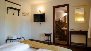 Habitación de hotel con cama, televisión montada en la pared, una mesita, un espejo y un baño visible a través de una puerta abierta.