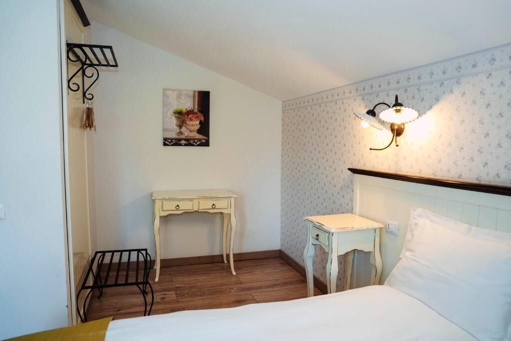 Un dormitorio con cama, escritorio, una mesita, lámpara de pared e imagen en la pared.