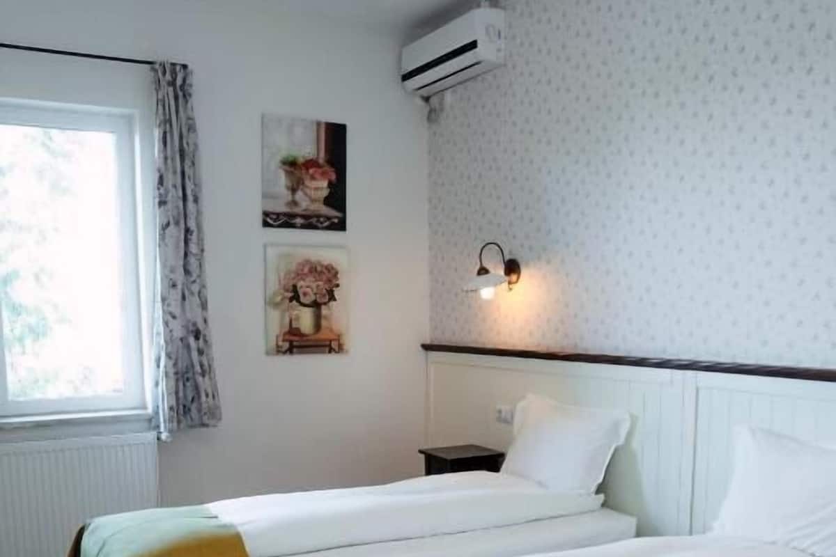 A hotel room with two beds, a window with curtains, an air conditioner, a nightstand, and wall-mounted lighting.