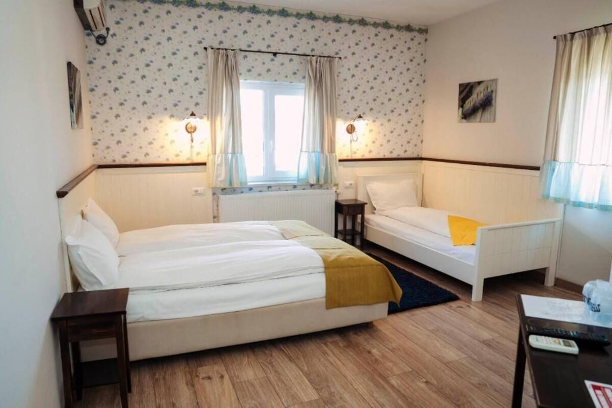 A room with two beds, a window with curtains, a radiator, a small table, and a wall-mounted light fixture.