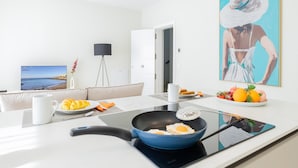 Uma cozinha moderna com uma frigideira no fogão, uma mesa de jantar com itens de café da manhã, uma televisão e um quadro na parede.