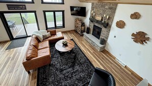 A modern living room with a leather sofa, a round coffee table, a stone fireplace, and large windows overlooking a deck. - Woods + Water Retreat (Lac du Flambeau)