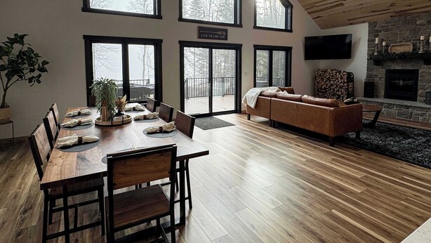 A spacious living room with a wooden ceiling, large windows, a stone fireplace, and a cozy seating area. - Woods + Water Retreat (Lac du Flambeau)