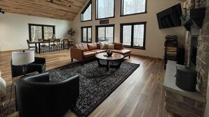 A spacious living room with a high ceiling, large windows, a fireplace, and a dining area. - Woods + Water Retreat (Lac du Flambeau)