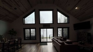 A spacious living room with a wooden floor, a large sofa, and a dining area with chairs and a table. There are multiple large windows providing natural light and views of the outdoors. - Woods + Water Retreat (Lac du Flambeau)