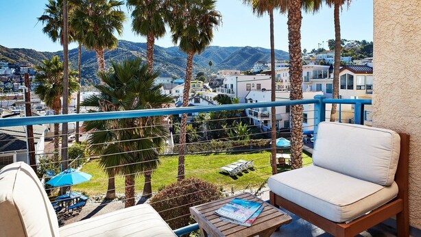 A balcony with a white sofa, a wooden table, and a magazine. There are palm trees and a view of a town and mountains in the background. - Charming Hideaway: 3 Family-Friendly Rooms, Near Catalina Island Museum (Avalon)