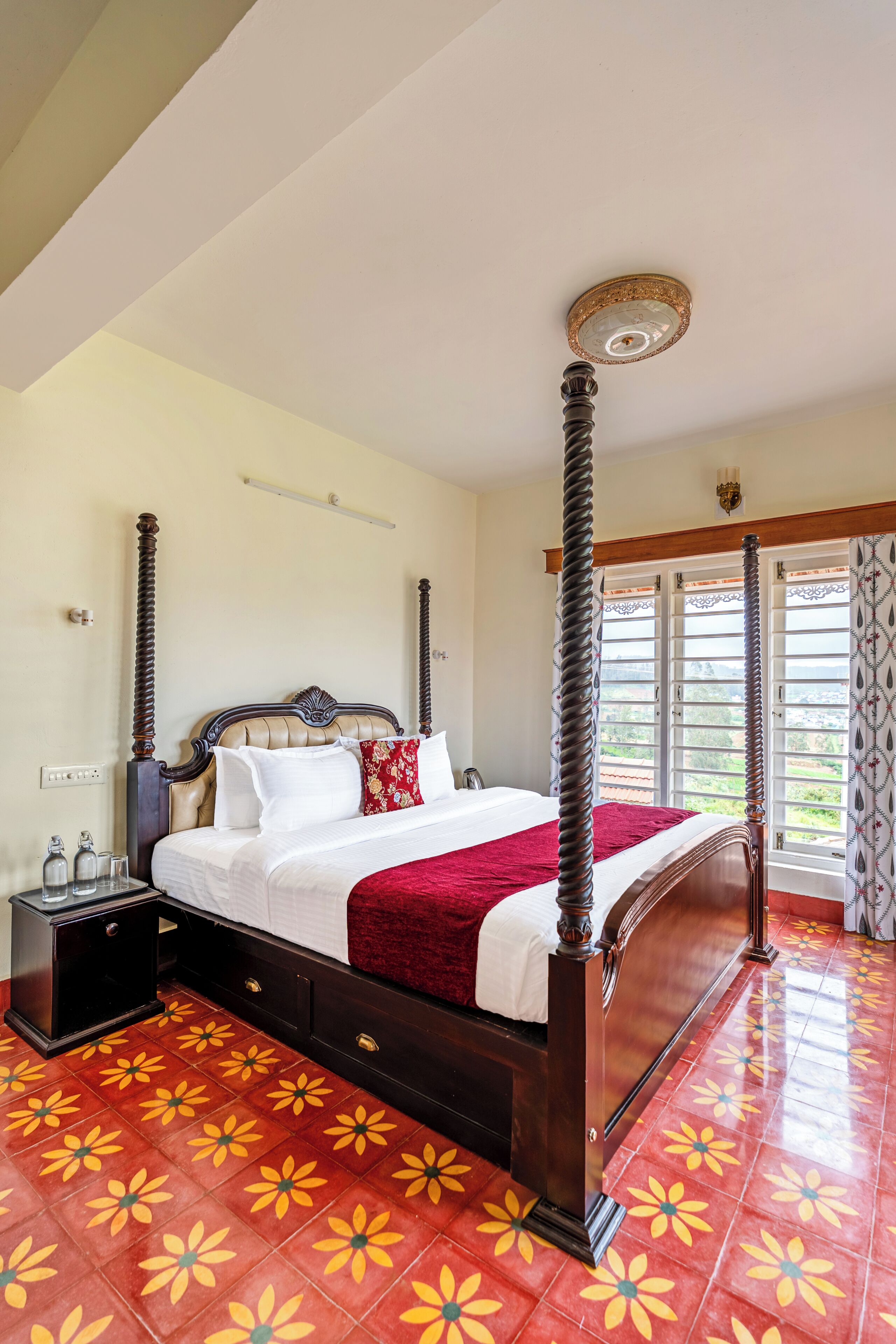A four-poster bed with a red bedspread, a nightstand with a lamp, and a window with patterned curtains.
