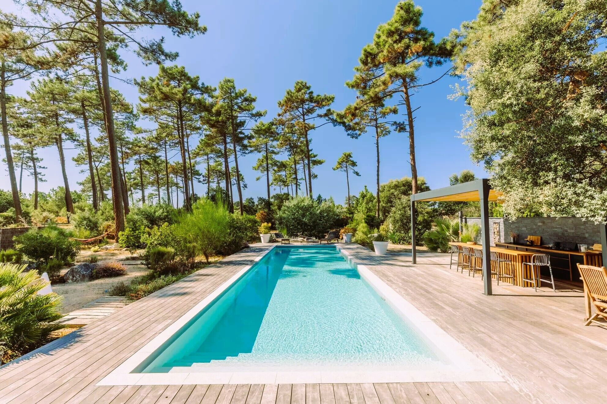 Una piscina rectangular con una cubierta de madera, rodeada de altos pinos y un muro de piedra.