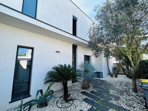 A modern building with a white facade, large windows, and a small garden area with plants and a tree. - Spacious modern villa with garage (La Grande-Motte)