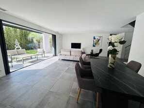 A modern living room with a dining area, a TV, and a large window overlooking a garden. - Spacious modern villa with garage (La Grande-Motte)