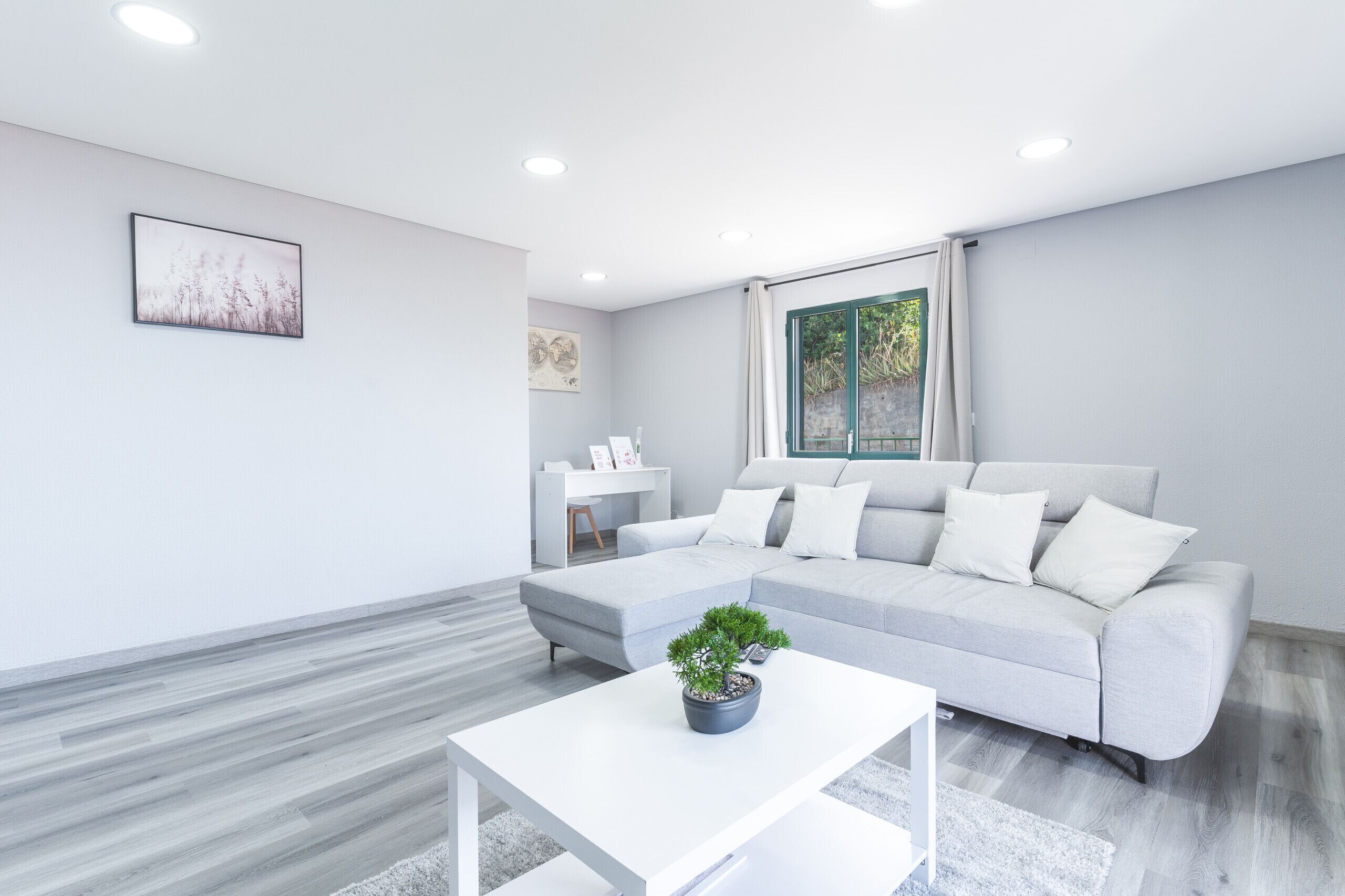 A modern living room with a grey sofa, a white coffee table with a plant, and a large window with curtains.
