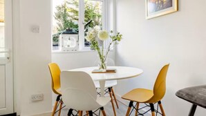 Un área de comedor con una mesa redonda, cuatro sillas, un florero con flores, un cuadro en la pared y un tragaluz.