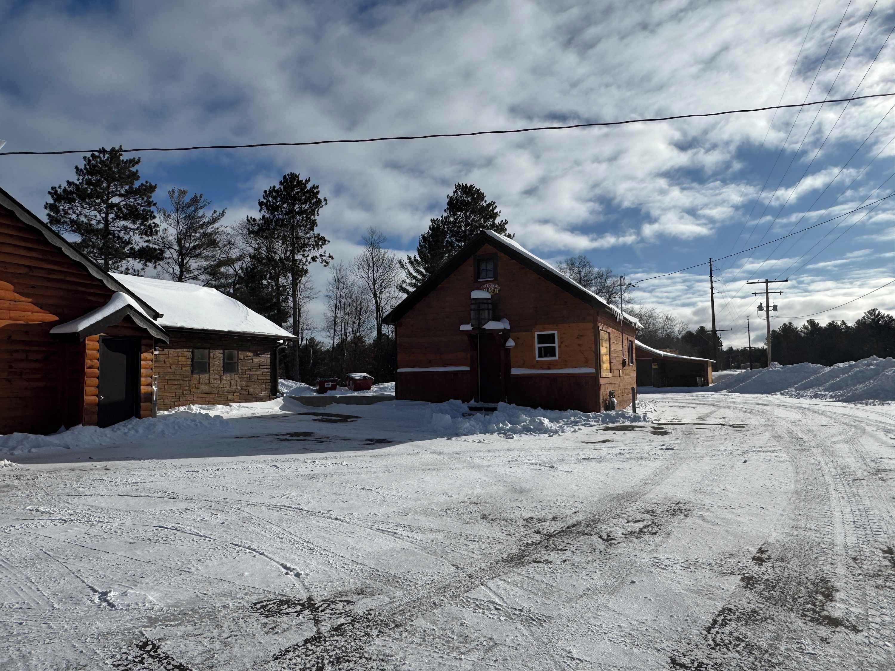 A wooden cabin with a snow-covered roof, a brick building adjacent to it, and a dirt road in the foreground.