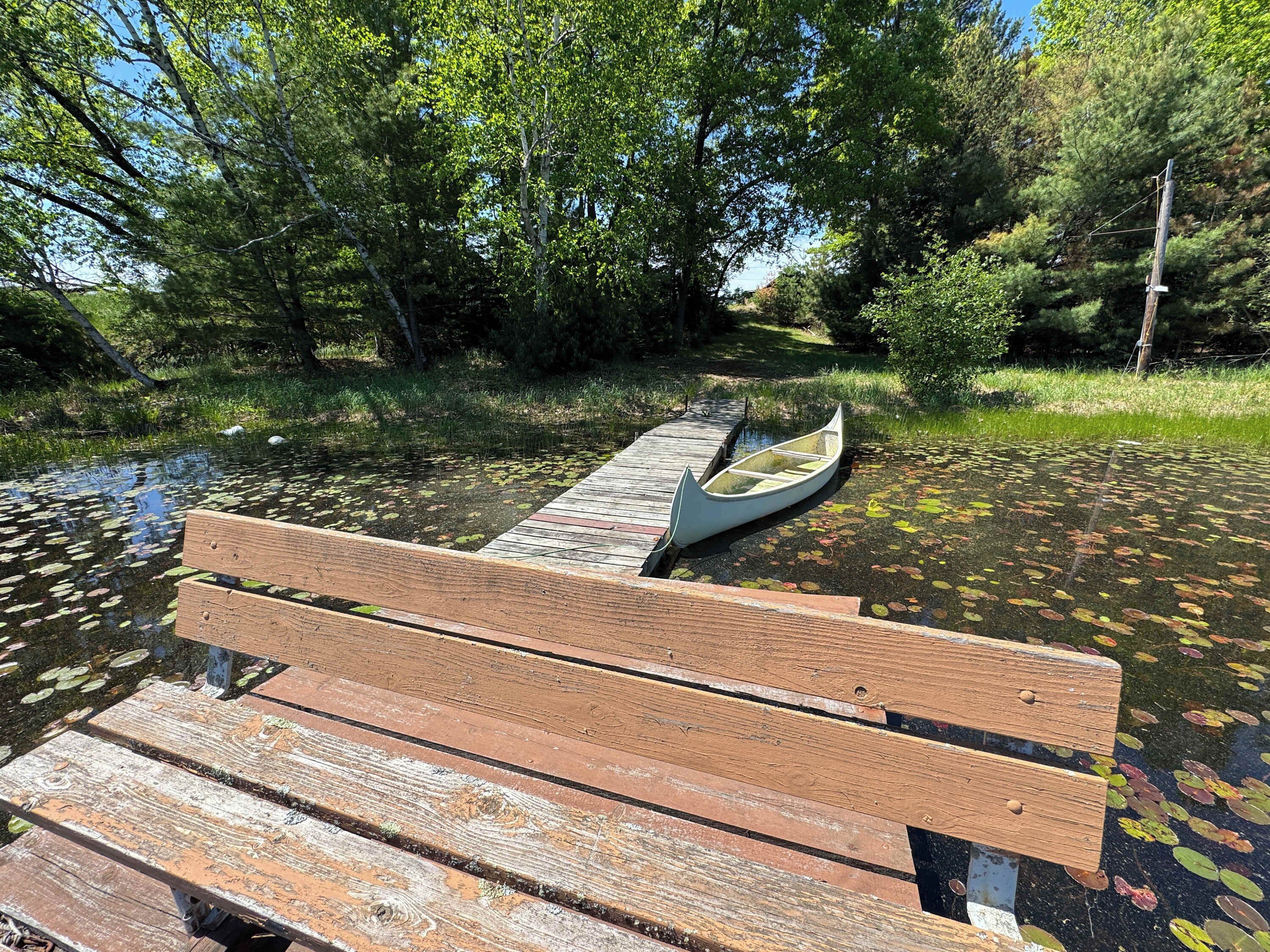 A wooden bench by a calm body of water with a canoe on a dock.