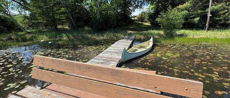 A wooden bench by a calm body of water with a canoe on a dock.
