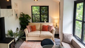 A living room with a beige sofa, a glass coffee table, a black chair, a large plant, and a window with a view of greenery. - Tranquil cottage stay with fireplace and pond (Oswestry)