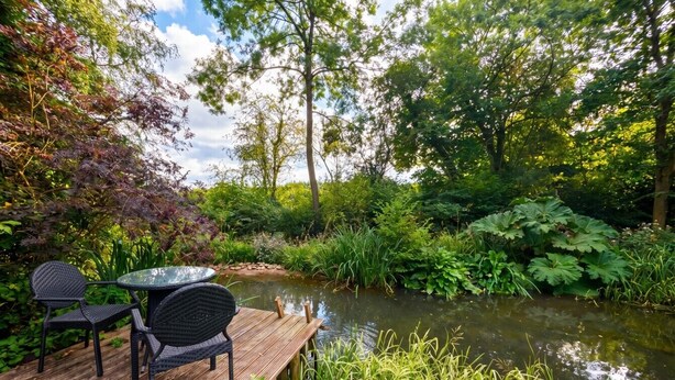 A wooden deck with a table and chairs overlooking a pond surrounded by lush greenery. - Tranquil cottage stay with fireplace and pond (Oswestry)