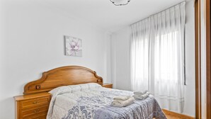 A bedroom with a wooden bed, a bedside table, a nightstand, a wall-mounted picture, and a window with sheer curtains. - Apartment 'Carmina El Fondon 3' with Balcony and Wi-Fi (Cangas de Onís)