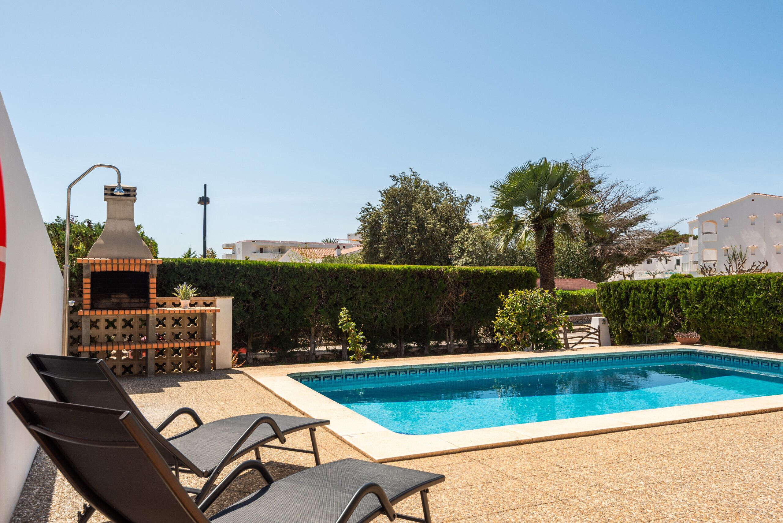 Una piscina con sillones de descanso, un horno de ladrillo y un seto.