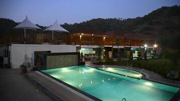 Una piscina con zona de estar hundida, un área de comedor al aire libre techada y un edificio de varios niveles con balcones.