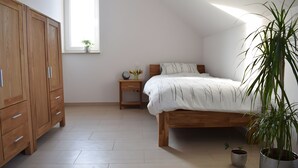 Ein Schlafzimmer mit einem hölzernen Bett, einem Nachttisch, einem Kleiderschrank, einem Fenster mit einer Pflanze und einem Blumentopf.