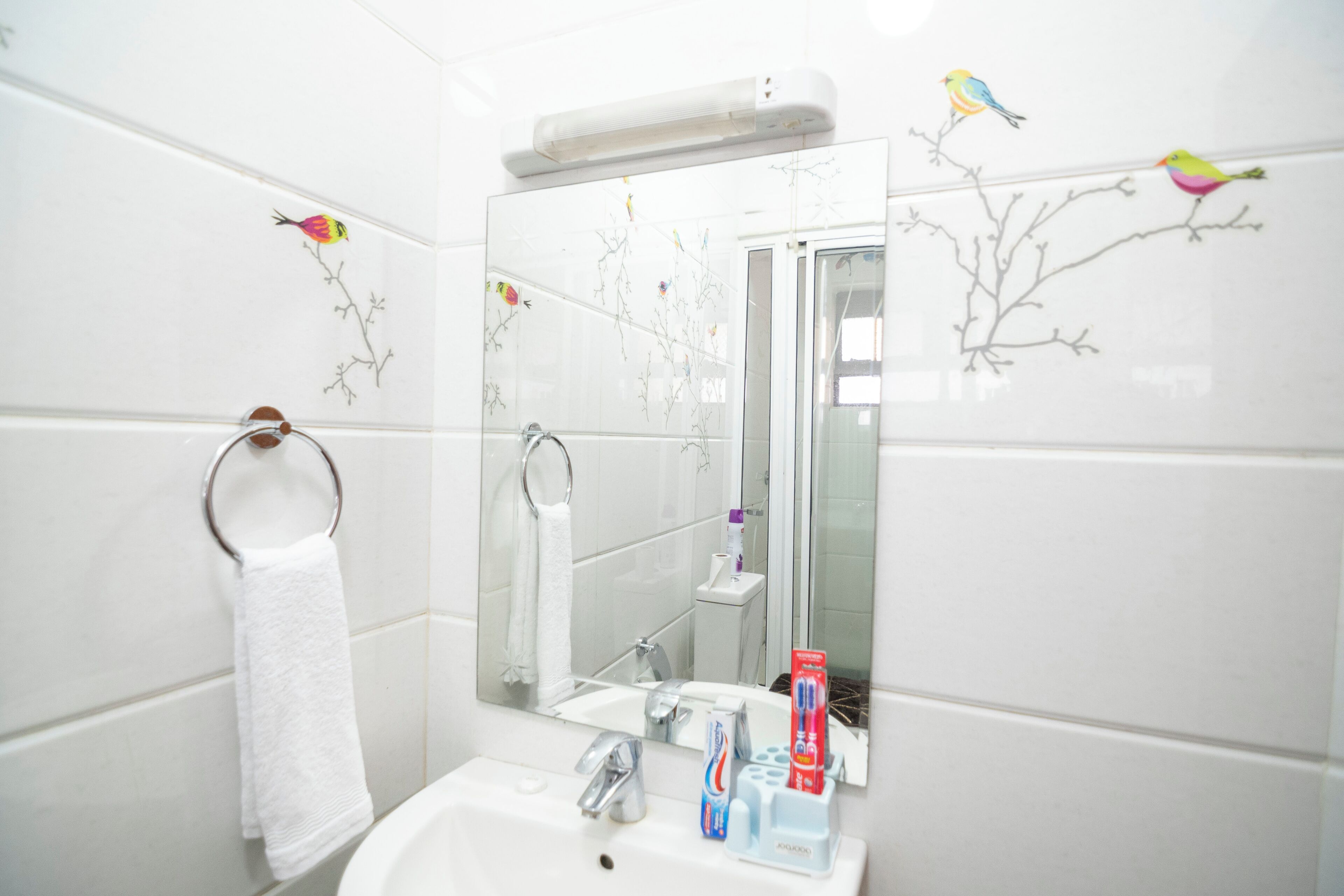 Een badkamer met witte tegels, een witte wastafel, een spiegel, een handdoekhouder en een tandenborstelhoudertje met tandpasta.